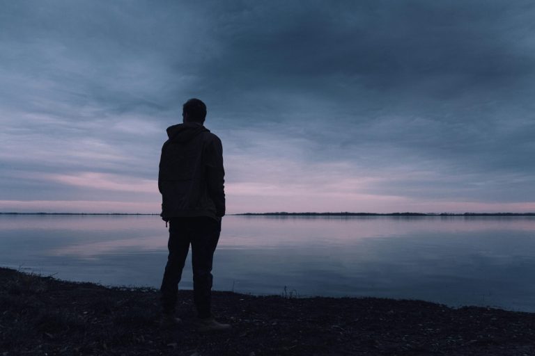 Silhouette of man working on addiction looking over calm lake with slight twilight with purple and blue hues.