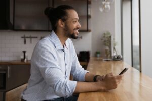 Man in his kitchen looking out window and at his phone for online rehab.